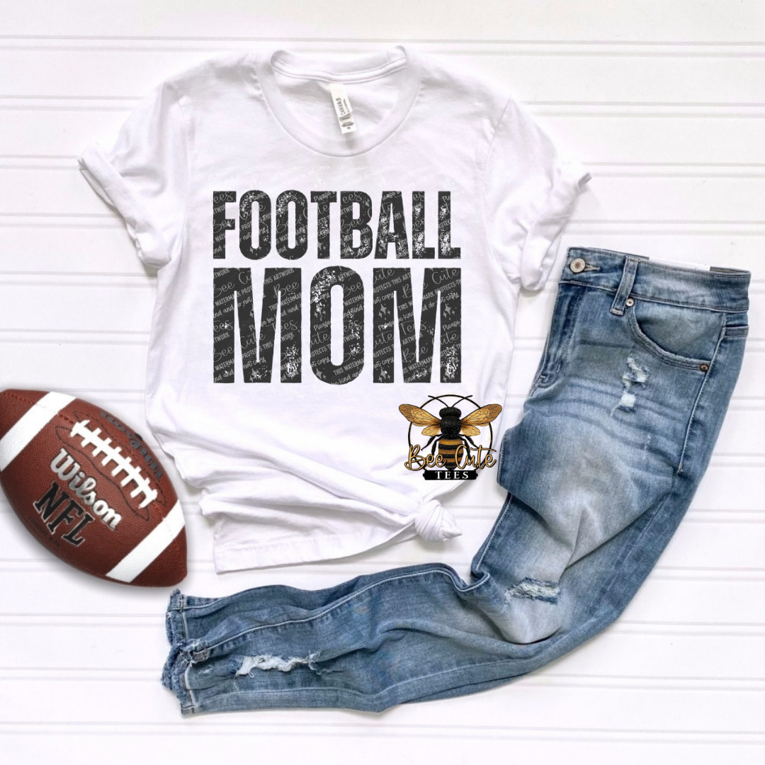 A white graphic t-shirt with 'FOOTBALL MOM' printed in distressed black letters, styled in a flat lay with blue jeans, a brown football, and a bee graphic on a white wood plank background.