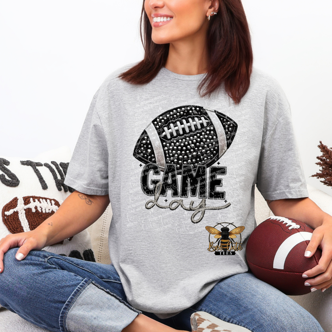 A woman wearing a heather gray graphic t-shirt with a black and white football design and the words 'GAME Day' in script. She is sitting cross-legged holding a brown football, with a matching decorative pillow in the background.