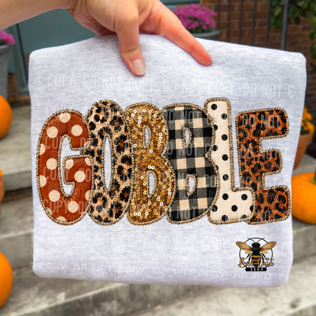 A folded light heather gray sweatshirt held by a hand, featuring the word 'GOBBLE' in large decorative letters with different patterned fills (polka dots, leopard print, sequins, buffalo check, polka dot) and a bee graphic at the bottom right.