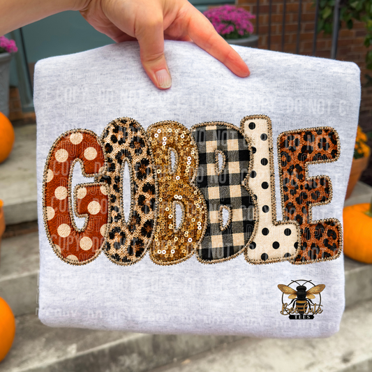 A folded light heather gray sweatshirt held by a hand, featuring the word 'GOBBLE' in large decorative letters with different patterned fills (polka dots, leopard print, sequins, buffalo check, polka dot) and a bee graphic at the bottom right.