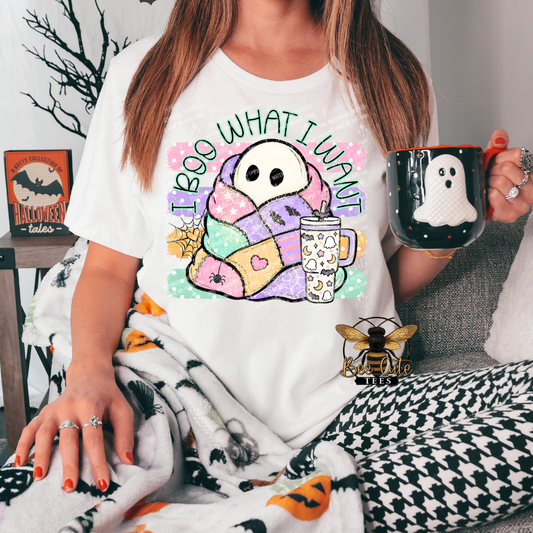 A person wearing a white graphic T-shirt with a pastel ghost design and the text 'I BOO WHAT I WANT' while holding a black mug with a ghost graphic. The scene is styled in a cozy indoor setting with a patterned blanket and Halloween-themed decor.