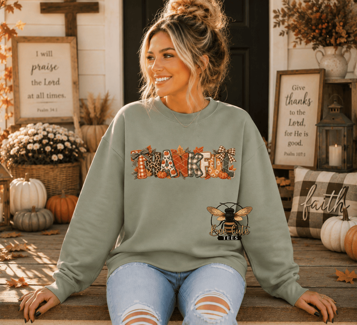 A woman sits on a wooden porch wearing a sage green sweatshirt with a 'THANKFUL' autumn-themed graphic and a bee logo. The background features fall decorations including pumpkins, a cross, and framed scripture.
