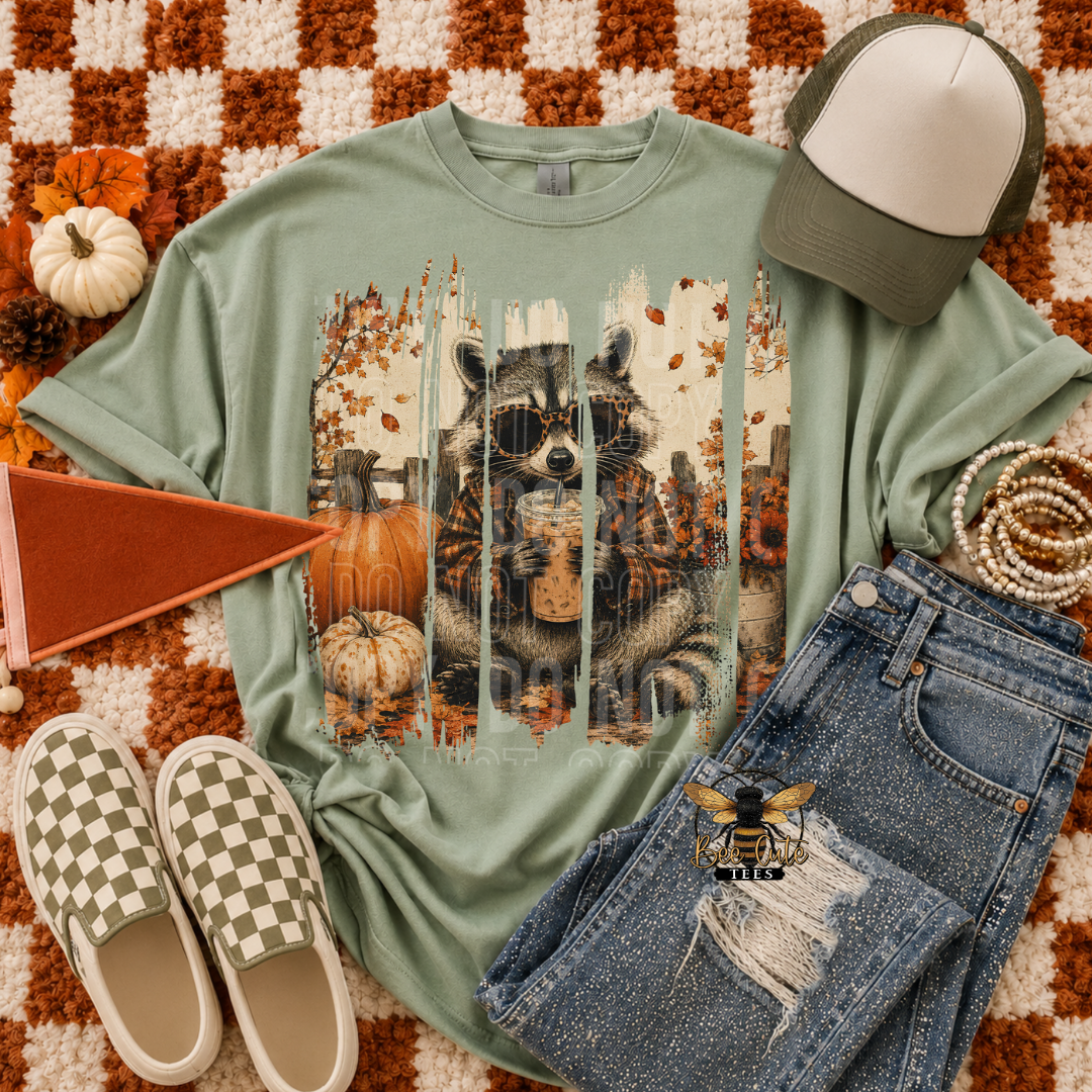 A styled flat-lay featuring a sage green graphic t-shirt with a raccoon and autumn print, paired with distressed blue jeans, checkered slip-on shoes, a trucker cap, and a small orange flag on a checkered rug.