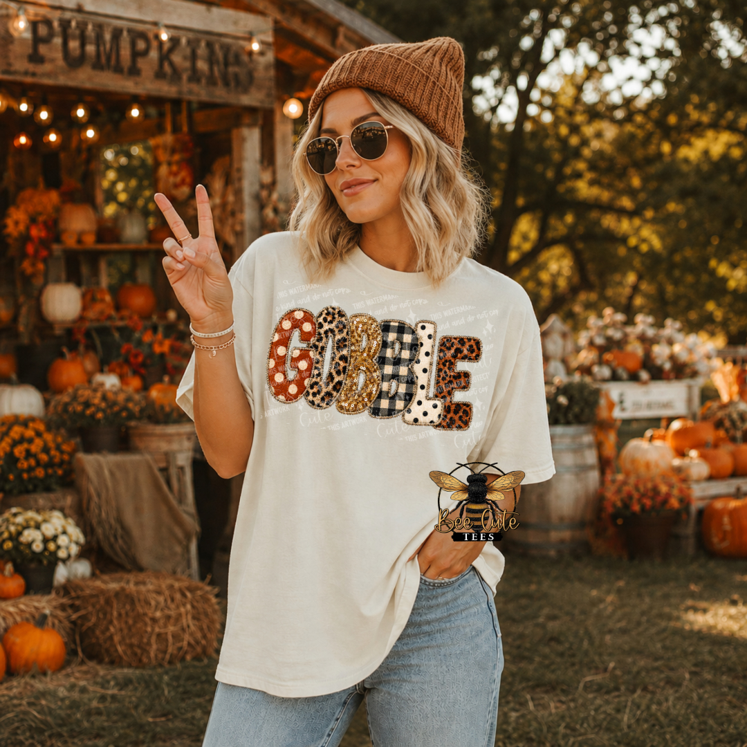 A woman wearing a cream graphic t-shirt with leopard-print applique spelling 'COOLE' stands in front of a rustic autumn pumpkin patch. She is wearing a brown knit beanie, sunglasses, and light-wash jeans, making a peace sign with one hand.