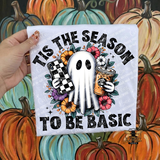 A hand holds a white graphic T-shirt featuring a ghost holding a checkered phone and a iced coffee, surrounded by flowers and the text 'TIS THE SEASON TO BE BASIC'. The background is a painted autumn pumpkin scene.