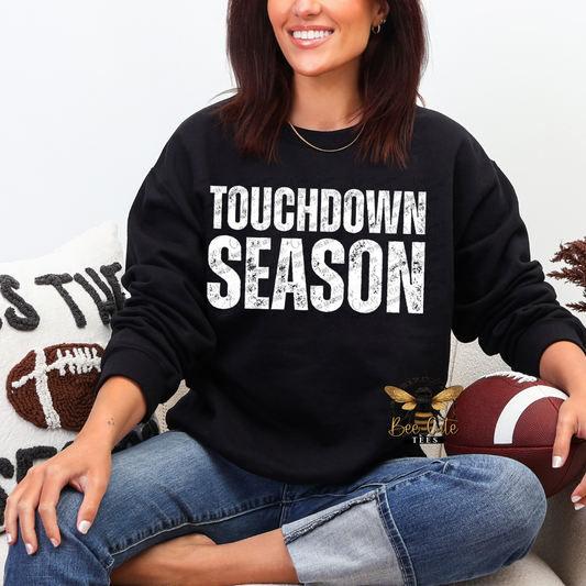 A woman sitting cross-legged wearing a black sweatshirt with 'TOUCHDOWN SEASON' printed in distressed white letters. She is holding a football, with a white throw pillow featuring a football graphic in the background.