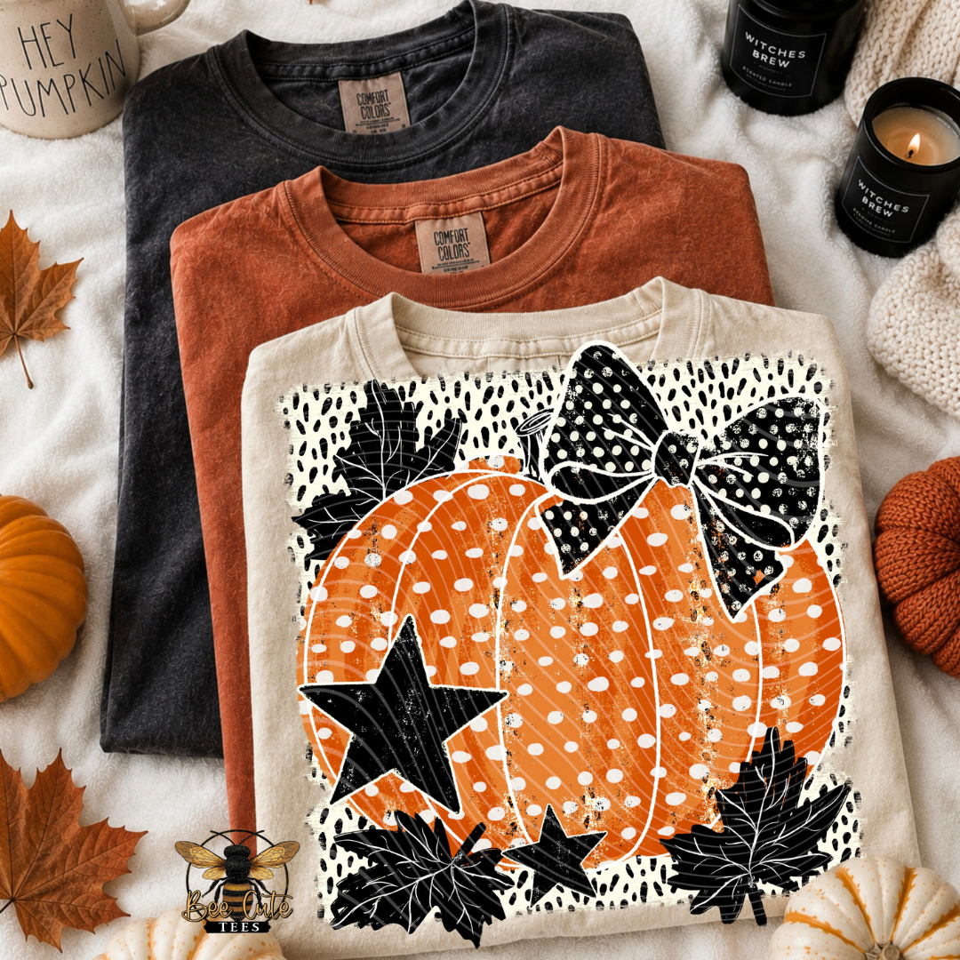 Three folded graphic t-shirts in dark charcoal, rust orange, and cream are arranged in a flat lay with autumn props including pumpkins, leaves, candles, and a mug.