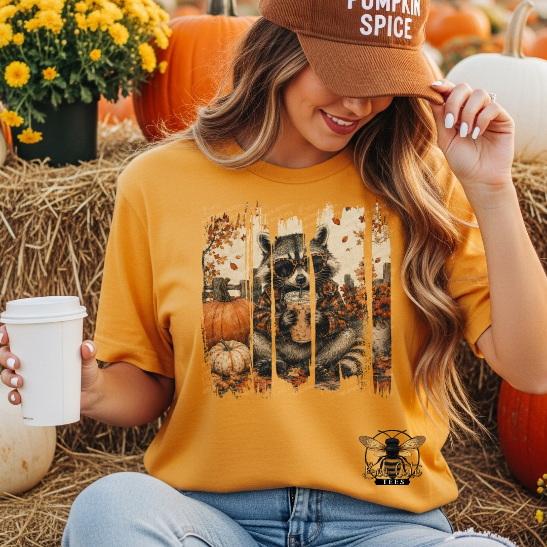 A woman wearing a mustard yellow graphic t-shirt with a fall-themed dog print, sitting in a pumpkin patch setting and holding a white coffee cup.
