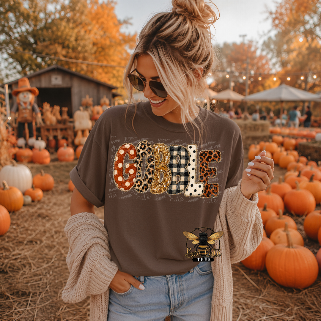 A woman wearing a brown graphic t-shirt with 'GOBBLE' spelled out in leopard print, gingham, and polka dot patterns, styled with a beige knit cardigan and sunglasses in a pumpkin patch.