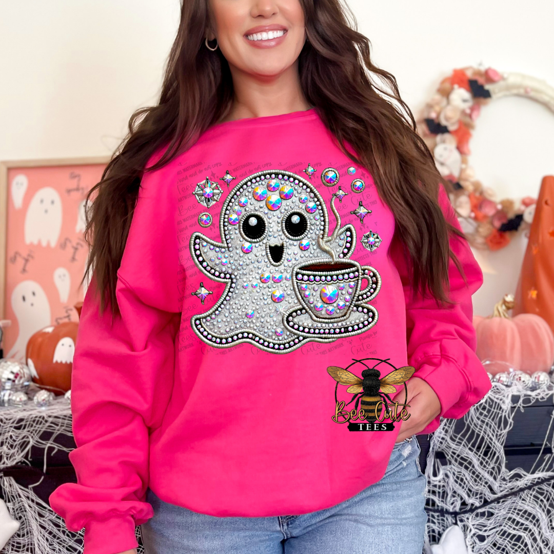 A woman wearing a bright pink crewneck sweatshirt with a large glittery ghost and coffee cup graphic on the front, paired with blue jeans in a Halloween-themed indoor setting.