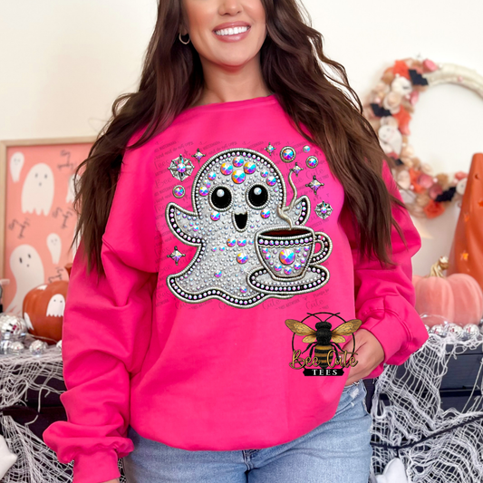 A woman wearing a bright pink crewneck sweatshirt with a large glittery ghost and coffee cup graphic on the front, paired with blue jeans in a Halloween-themed indoor setting.