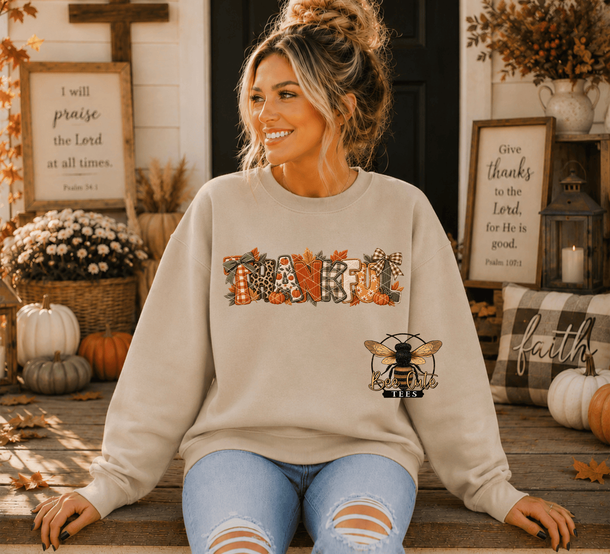 A woman sitting on a wooden porch wearing a beige crewneck sweatshirt with a colorful 'Thanksgiving' graphic and a bee logo on the front, paired with distressed light blue jeans.