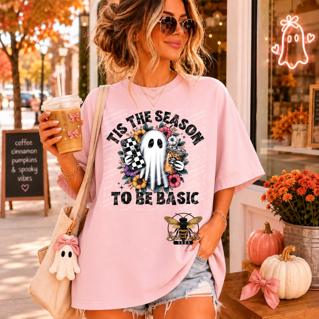 A woman wearing a light pink graphic t-shirt with a ghost and bee logo that reads 'TIS THE SEASON TO BE BASIC', holding a coffee cup in a cafe setting.
