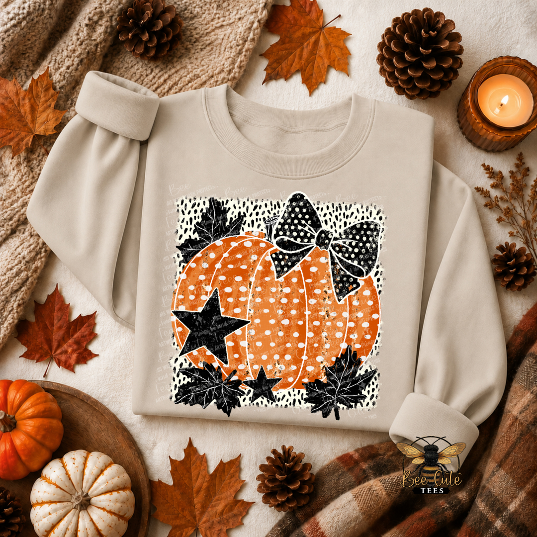 A beige long-sleeve sweatshirt laid flat on a cozy autumn-themed background with pinecones, fall leaves, a small pumpkin, plaid fabric, and a lit candle. The sweatshirt features a square graphic print of an orange pumpkin with white polka dots, black bows, and black stars.