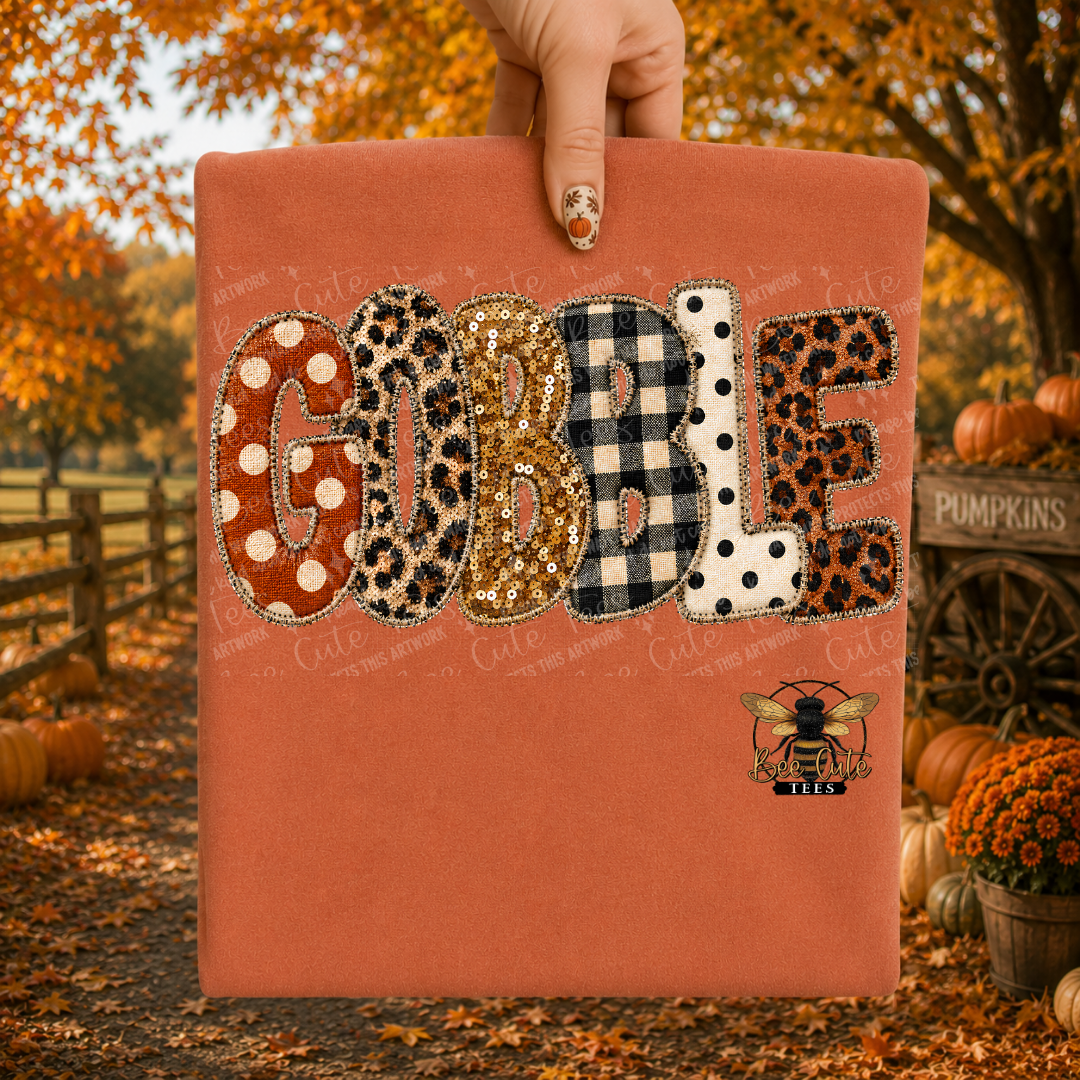 A folded terracotta orange t-shirt is held up by a hand against an autumn outdoor background with pumpkins, fall leaves, and trees. The shirt features the word 'GOBBLE' in large applique-style letters with various patterns including leopard print, gingham, polka dots, and glitter, along with a small bee logo.