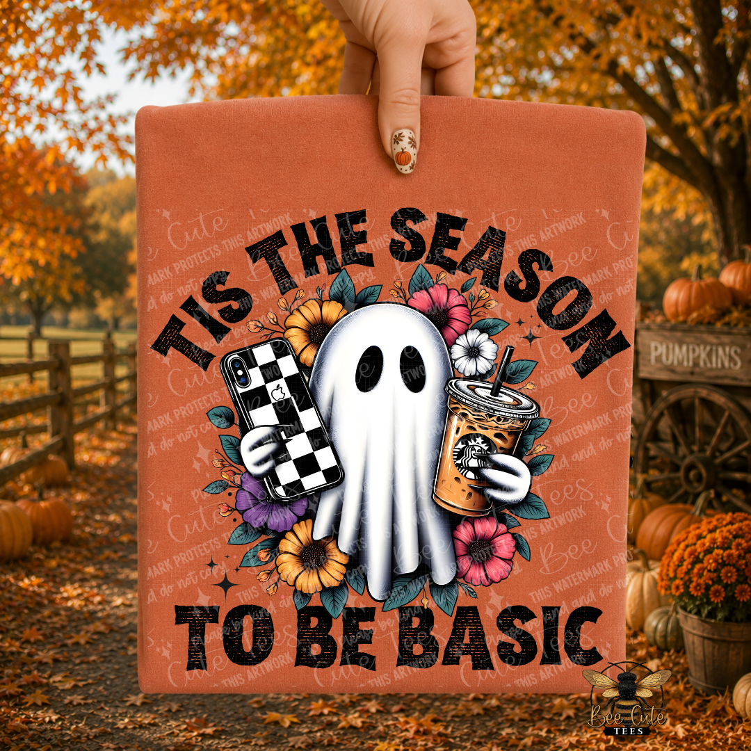 A hand holds up a rust-orange graphic t-shirt featuring a white cartoon ghost holding a black-and-white checkered phone case and a coffee drink, surrounded by flowers, with the text 'TIS THE SEASON TO BE BASIC'. The background is a blurred autumn outdoor scene with pumpkins and fall foliage.