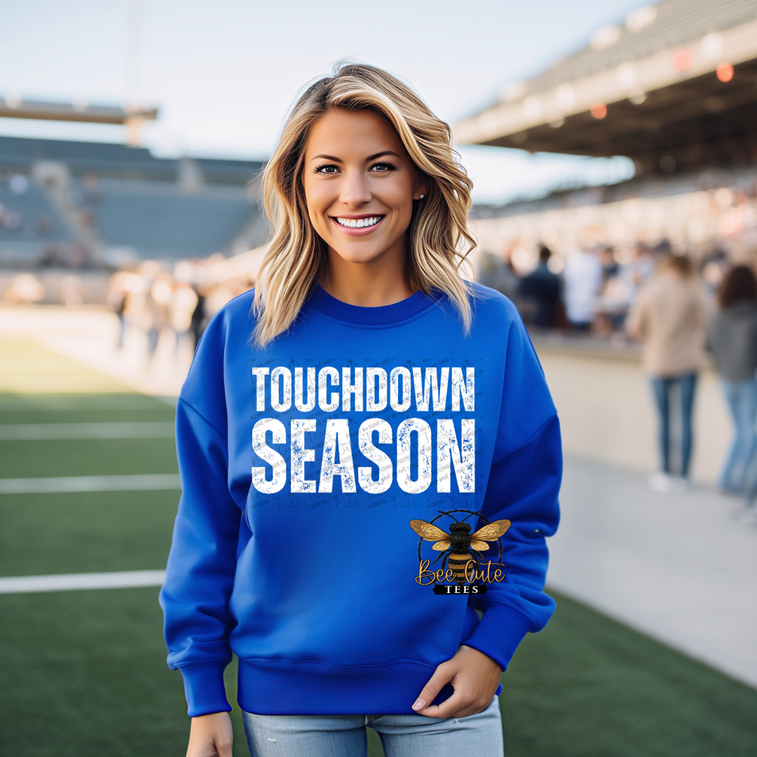 A smiling woman stands on a football field wearing a bright royal blue sweatshirt with 'TOUCHDOWN SEASON' printed in distressed white letters across the chest, plus a bee logo near the lower right front. The background shows blurred stadium seating and field activity.