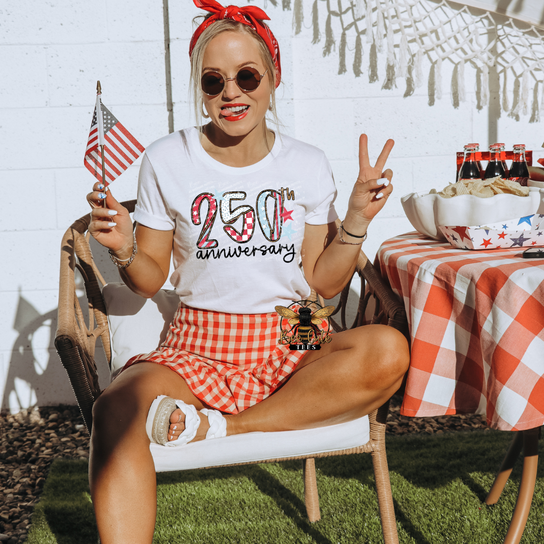 Woman in a casual outfit with a '250th anniversary' shirt, sitting outdoors with a table and American flags.
