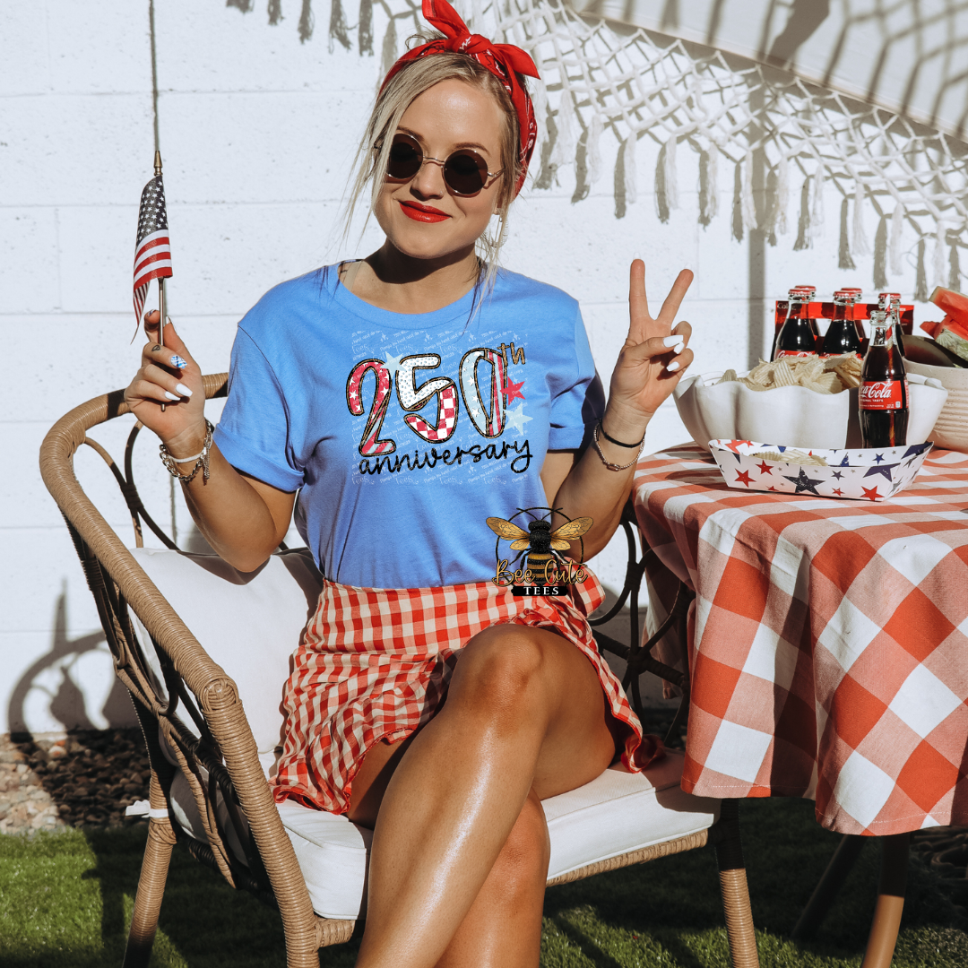Woman in a blue t-shirt with '250th anniversary' text, sunglasses, and red checkered skirt sitting outdoors with a table and bottles in the background.