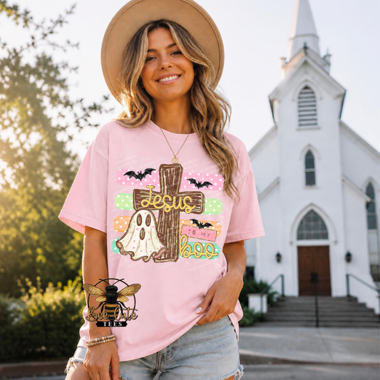 A woman wearing a light pink graphic t-shirt with a Halloween design featuring a cross, ghost, bats, and the words 'Jesus is my boo' stands in front of a white church.