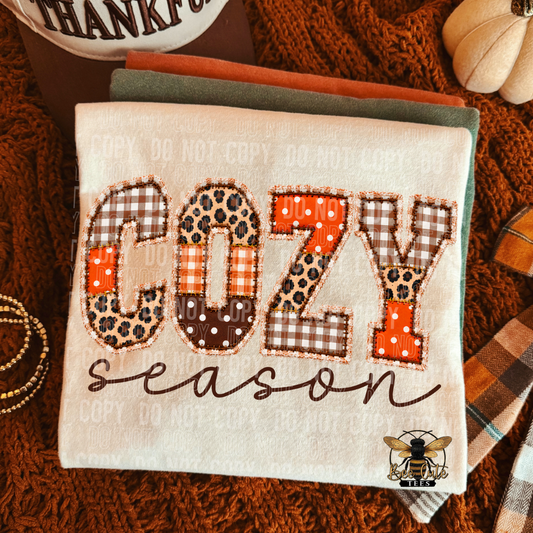A folded cream-colored t-shirt featuring the words 'COZY season' in decorative applique lettering with leopard print, gingham, and orange polka dot patterns, styled on a brown knit blanket with autumn props.