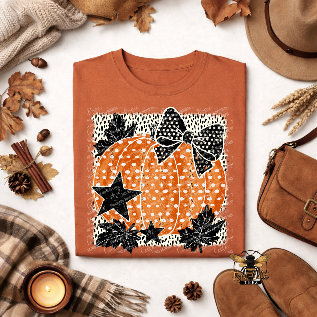 An orange folded t-shirt with a pumpkin graphic, styled in a flat lay with autumn props including a hat, scarf, candle, and shoes on a white background.