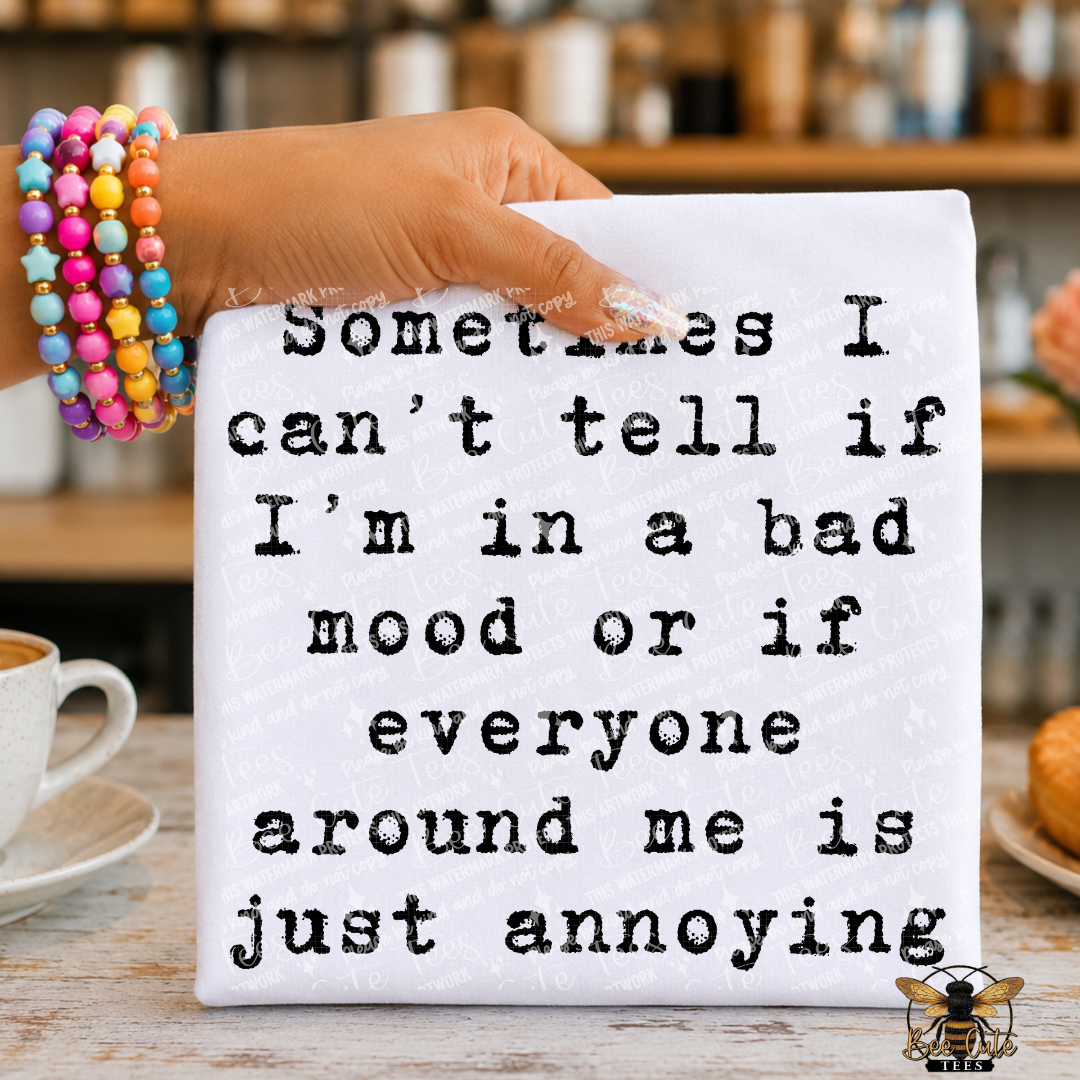 A hand holding a white t-shirt with black typewriter-style text that reads, 'Sometimes I can't tell if I'm in a bad mood or if everyone around me is just annoying.' The background shows a blurred kitchen or cafe setting with a cup, pastry, and a colorful beaded bracelet on the wrist.