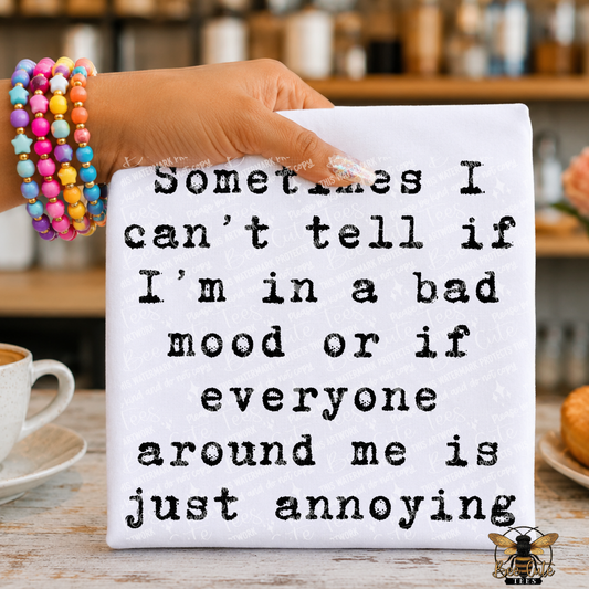A hand holding a white t-shirt with black typewriter-style text that reads, 'Sometimes I can't tell if I'm in a bad mood or if everyone around me is just annoying.' The background shows a blurred kitchen or cafe setting with a cup, pastry, and a colorful beaded bracelet on the wrist.