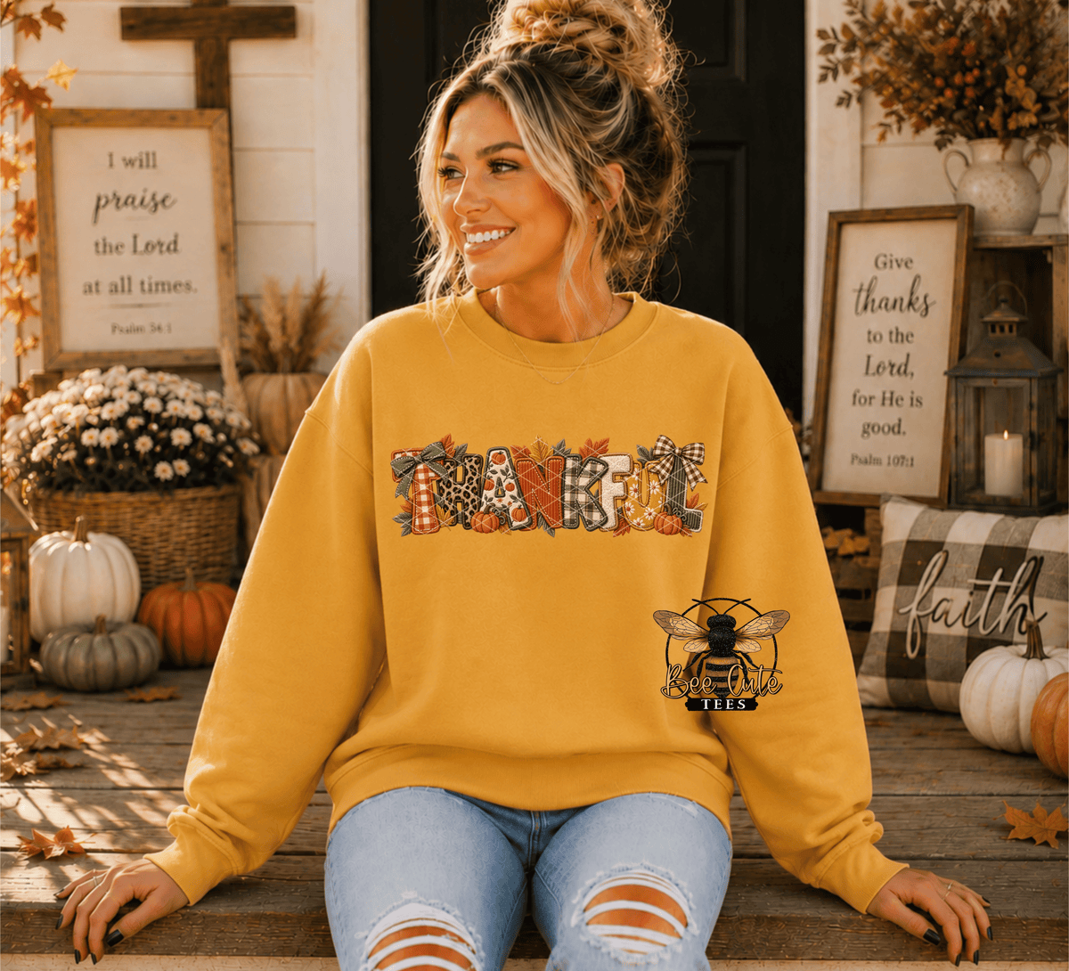 A woman wearing a mustard yellow crewneck sweatshirt with a 'THANKFUL' autumn-themed graphic and a bee logo, sitting on a wooden porch with fall decorations.