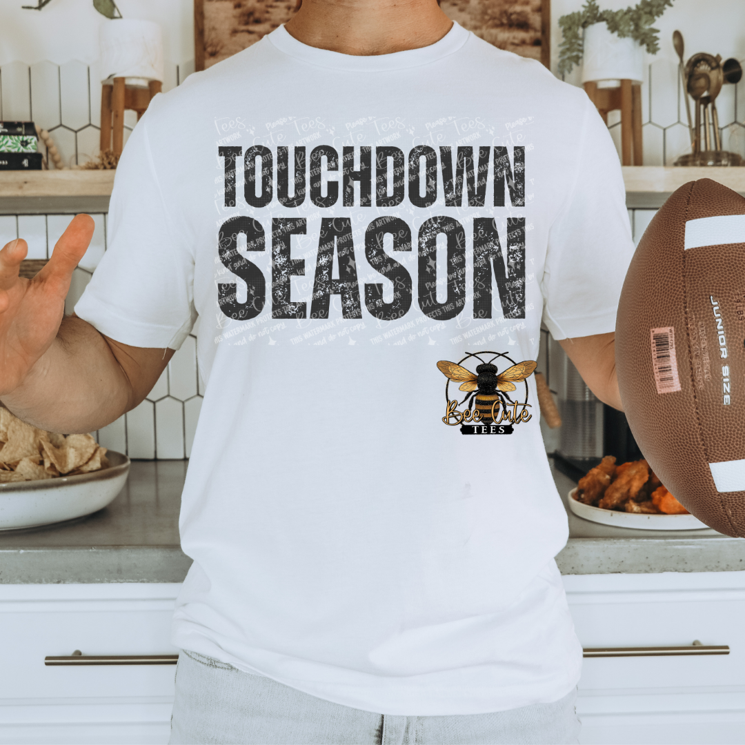 A person wearing a white graphic T-shirt with the words 'TOUCHDOWN SEASON' in distressed black letters and a bee logo below, holding a football in a kitchen setting.