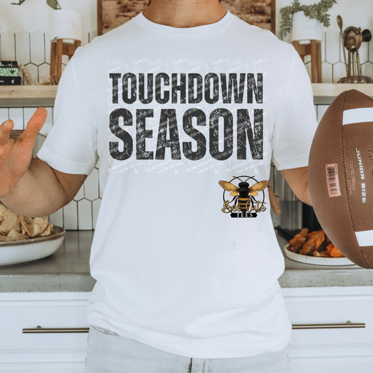 A person wearing a white graphic T-shirt with the words 'TOUCHDOWN SEASON' in distressed black letters and a bee logo below, holding a football in a kitchen setting.
