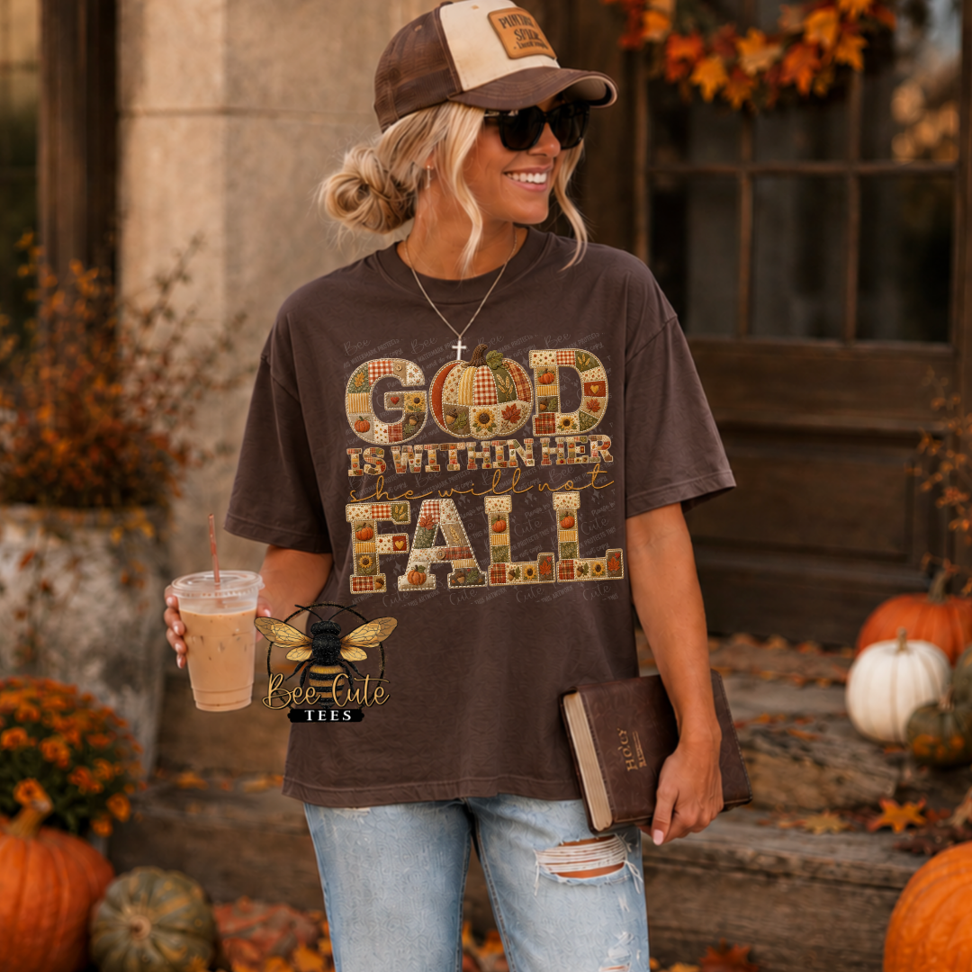 A woman wearing a brown graphic t-shirt with 'GOD IS WITHIN HER FALL' in a patchwork autumn design, styled with a brown and beige baseball cap, sunglasses, and ripped blue jeans. She is holding an iced coffee and a book in an outdoor autumn setting with pumpkins and fall leaves.