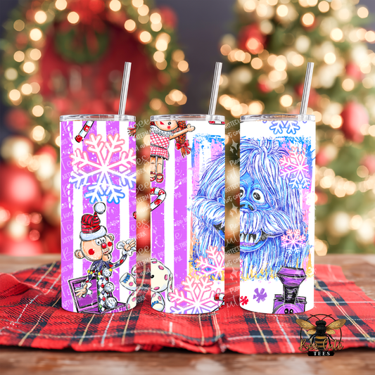 Three decorated skinny tumblers with metal straws are displayed on a red plaid blanket in front of a blurred Christmas tree and lights. The tumblers feature holiday illustrations including snowflakes, a Santa-clad character, a nutcracker, a dog, and a blue furry creature.