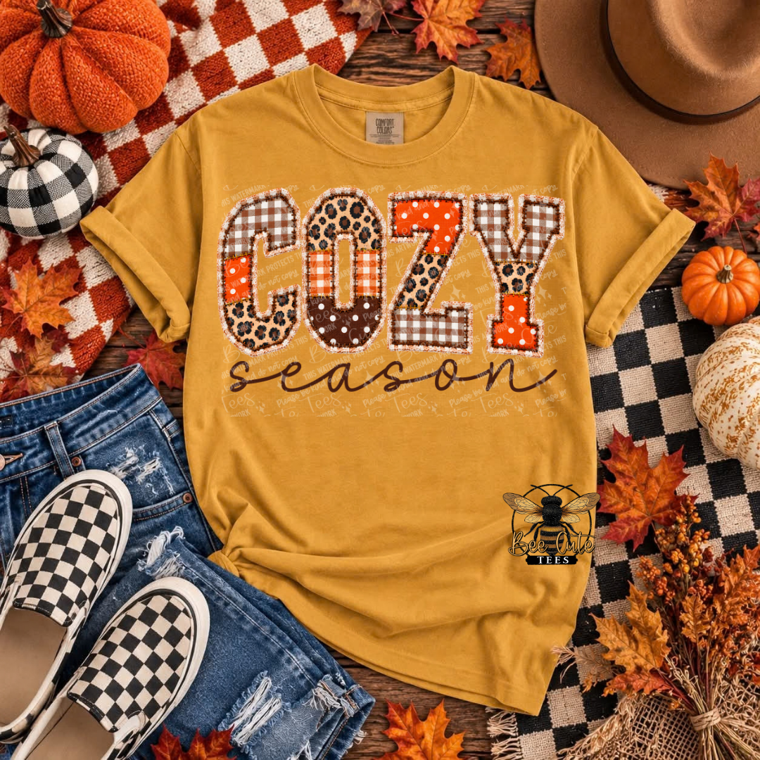A mustard yellow graphic T-shirt laid flat with a fall-themed styled arrangement. The shirt reads 'COZY season' in decorative patterned letters, with a small bee logo near the lower right front.