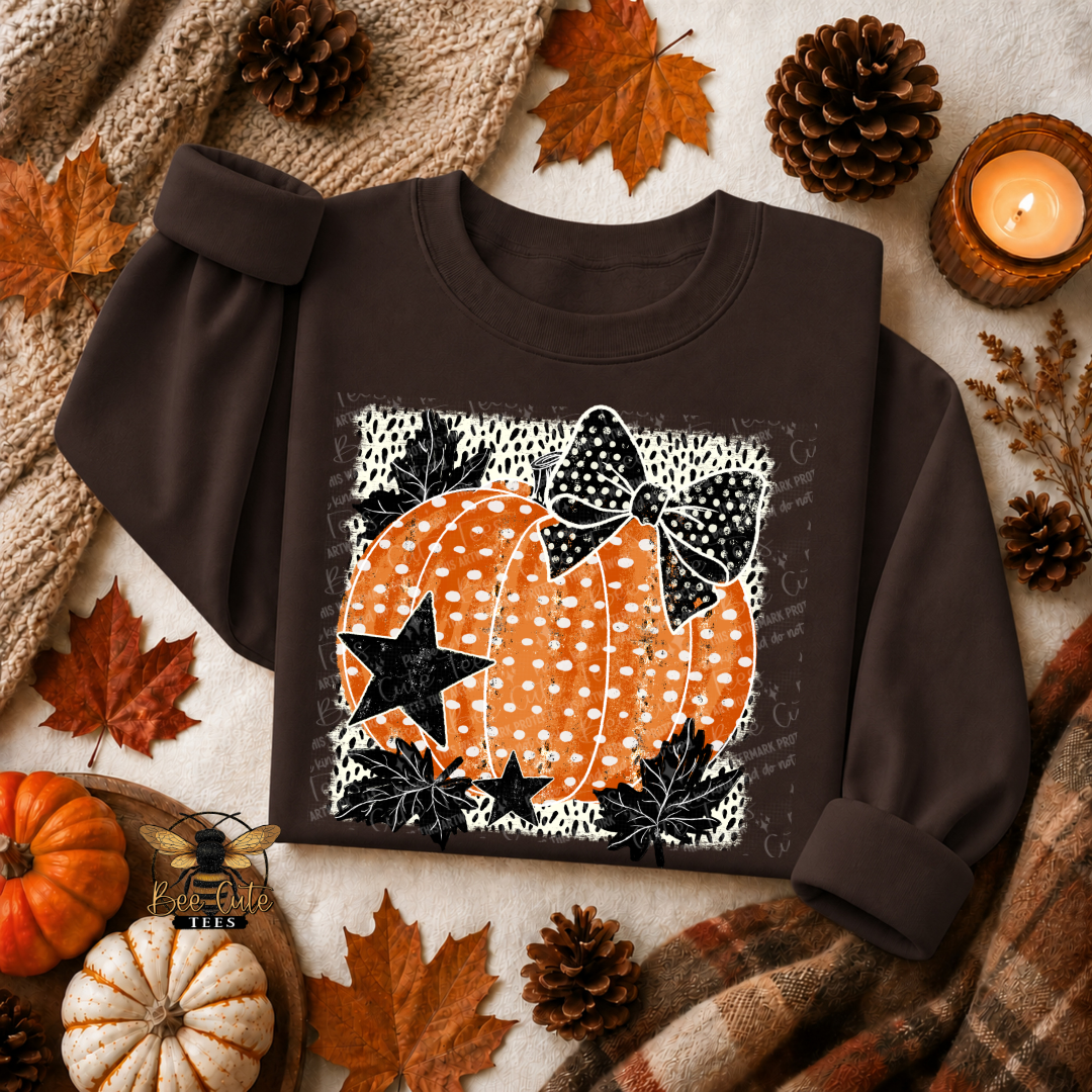 A dark brown long-sleeve sweatshirt laid flat on a neutral surface, surrounded by autumn props including pinecones, orange leaves, a small pumpkin, a candle, and a plaid scarf. The sweatshirt has a large front graphic showing two orange pumpkins with white polka dots, black stars, and a black bow on a white patterned background.