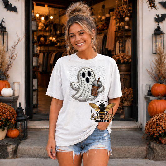 A woman stands in front of a storefront wearing an oversized white graphic T-shirt with a ghost holding a coffee cup design and denim shorts. The scene is decorated with pumpkins, fall foliage, and lanterns.