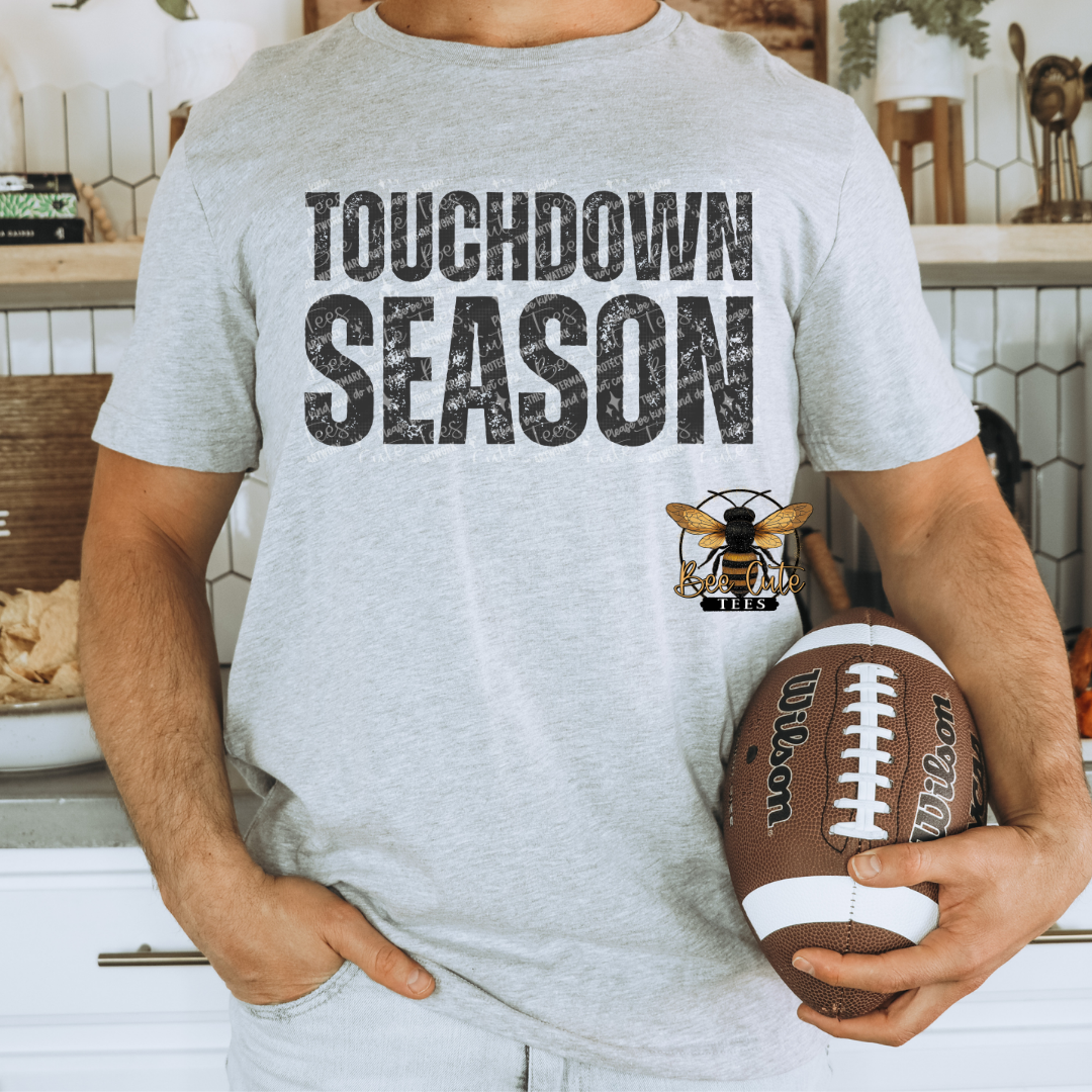A man wearing a light gray heather t-shirt with 'TOUCHDOWN SEASON' printed in distressed black letters and a bee logo, holding a  football in a kitchen setting.