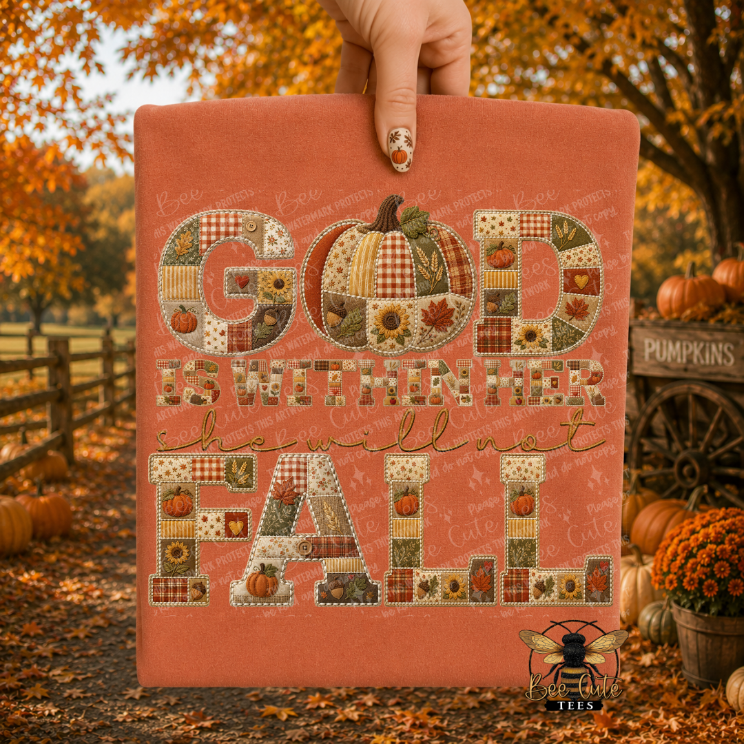 A hand holds up a rust-colored folded t-shirt with a patchwork autumn design that reads “GOD IS WITHIN HER she will not FALL.” The sign features patchwork-style letters and a pumpkin motif in fall colors, set against a blurred outdoor fall yard background with pumpkins and orange foliage.