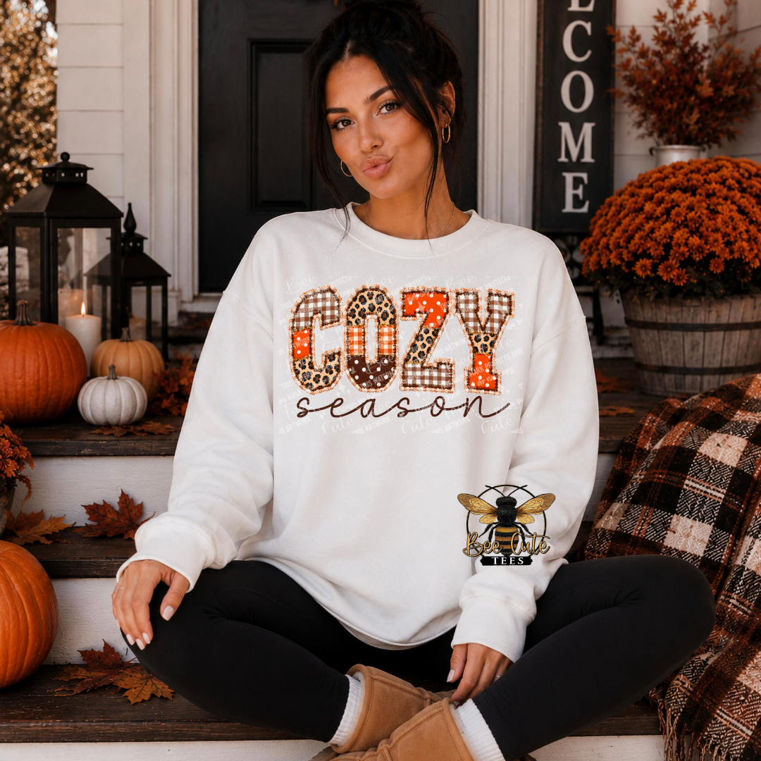 A woman sits cross-legged on a front porch wearing a white sweatshirt with 'COZY season' in a fall-themed patterned font. The word 'COZY' features leopard print, polka dots, and orange stripes, while 'season' is in black script.