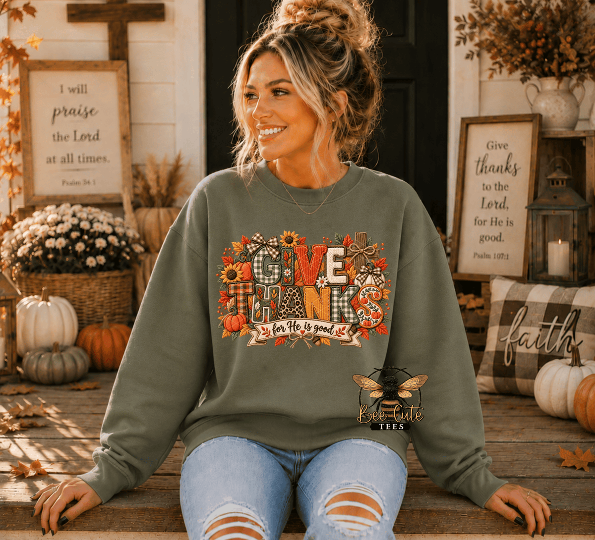 A woman wearing an olive green crewneck sweatshirt with a colorful 'Give Thanks' fall harvest graphic, sitting on a wooden porch decorated for autumn.