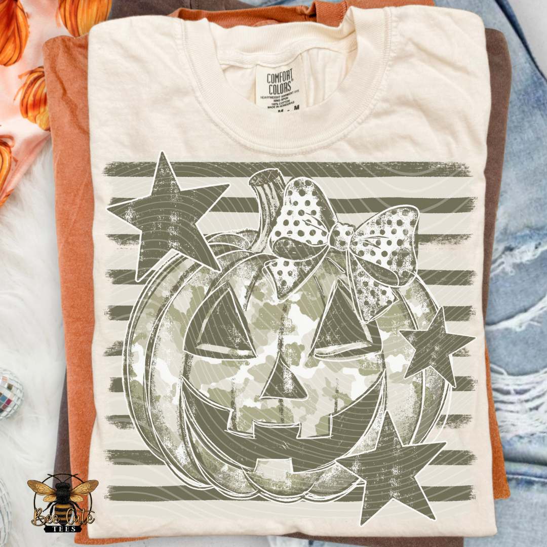 A folded cream-colored t-shirt with a Halloween graphic print, layered over an orange shirt and a blue denim shirt. The graphic features a camouflage pumpkin, a polka-dot bow, and stars in olive green on a striped background.