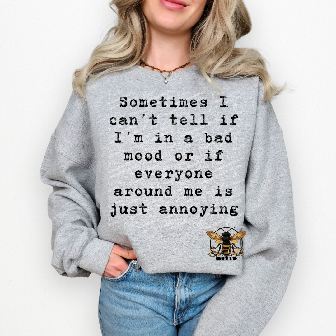 A woman wearing a light gray crewneck sweatshirt with black printed text that reads 'Sometimes I can't tell if I'm in a bad mood or if everyone around me is just annoying' and a small bee graphic near the lower right front. The sweatshirt is shown on a model against a plain white background.