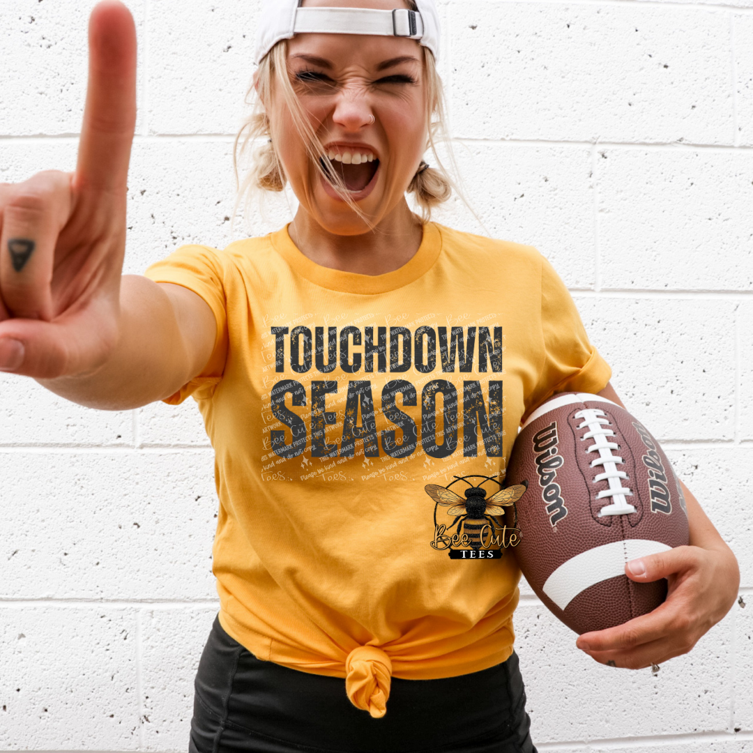 A woman wearing a yellow graphic t-shirt that reads 'TOUCHDOWN SEASON' with a bee logo, holding a football and making a peace sign against a white brick wall.