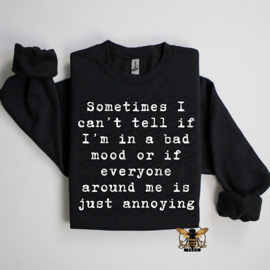 A black crewneck sweatshirt with white printed text reading 'Sometimes I can't tell if I'm in a bad mood or if everyone around me is just annoying' laid flat on a light gray background.