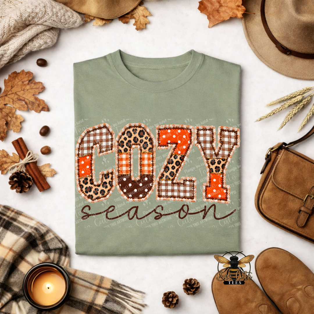 A folded sage green t-shirt with a 'COZY season' graphic in autumn-themed plaid and leopard print patterns, styled in a flat lay with fall accessories like a hat, scarf, candle, and shoes.