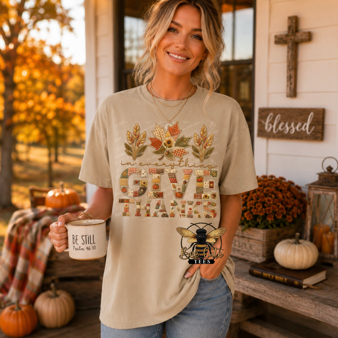 A woman stands on a decorated autumn porch wearing a beige graphic T-shirt with fall leaves and the words “Give Thanks,” holding a mug that says “Be Still” and a small decorative bee ornament.