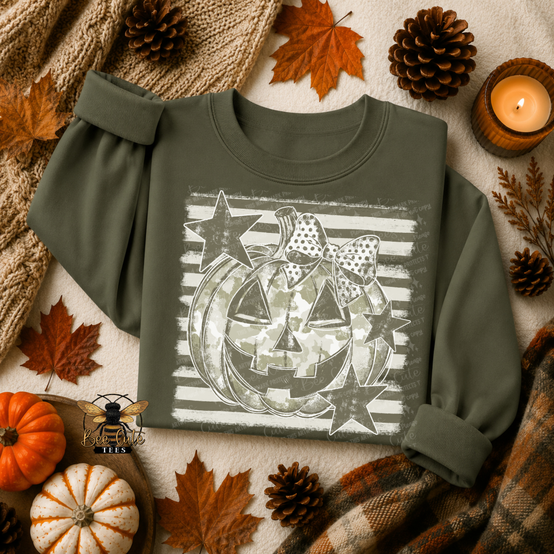 An olive green long-sleeve sweatshirt with a distressed Halloween graphic of a jack-o'-lantern, stars, and a bow, styled in a flat lay with autumn leaves, pumpkins, pinecones, and a candle.