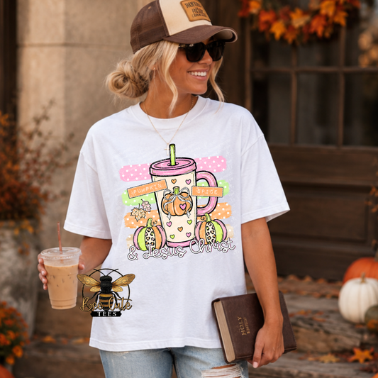 A woman wearing a white graphic t-shirt with a fall-themed pumpkin and latte art design, holding an iced coffee and a book in an outdoor autumn setting.