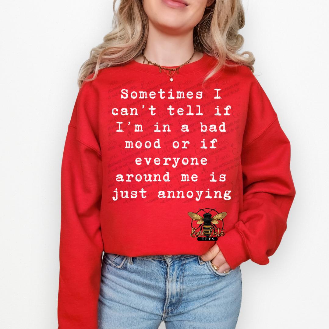 A woman wearing a bright red cropped sweatshirt with white text that reads, 'Sometimes I can't tell if I'm in a bad mood or if everyone around me is just annoying,' plus a small bee logo on the lower right chest. She is also wearing light blue jeans against a plain white background.