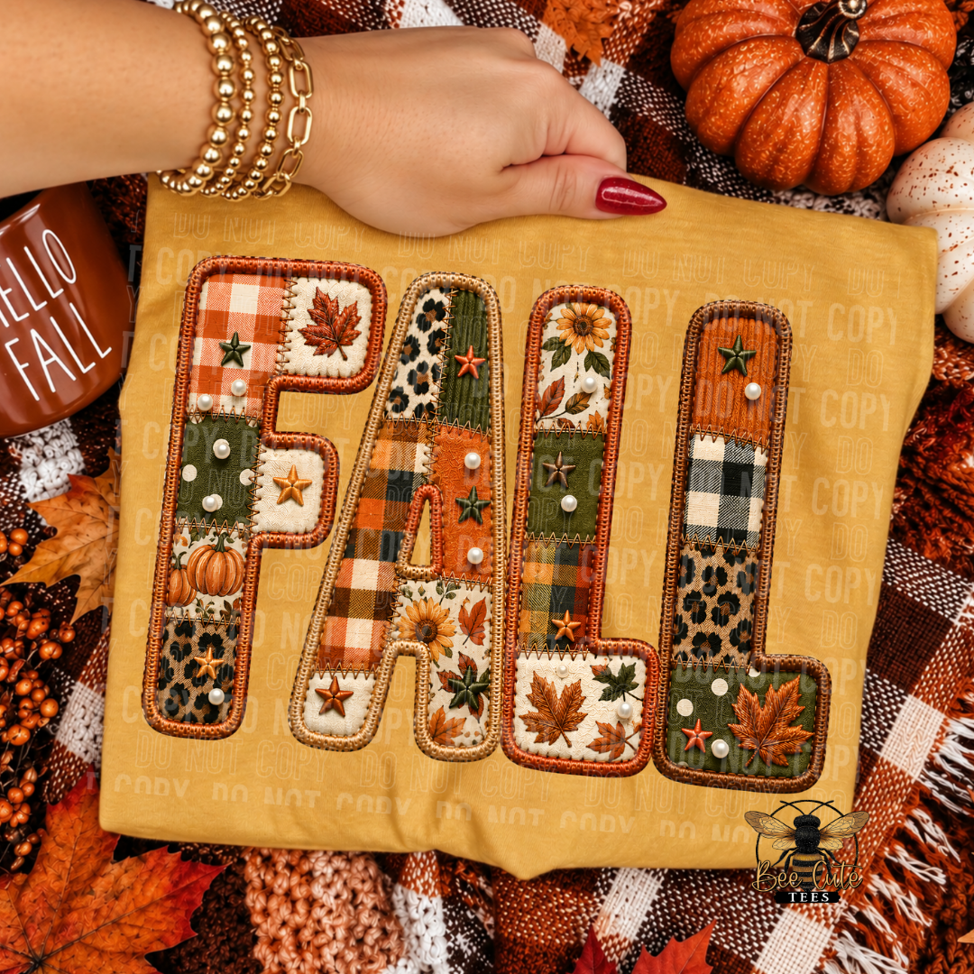 A close-up of a yellow folded t-shirt, featuring the word 'FALL' in large letters filled with an autumn patchwork design of plaid, leopard print, sunflowers, and leaves. A person's hand with red nail polish and gold bracelets rests on the fabric.